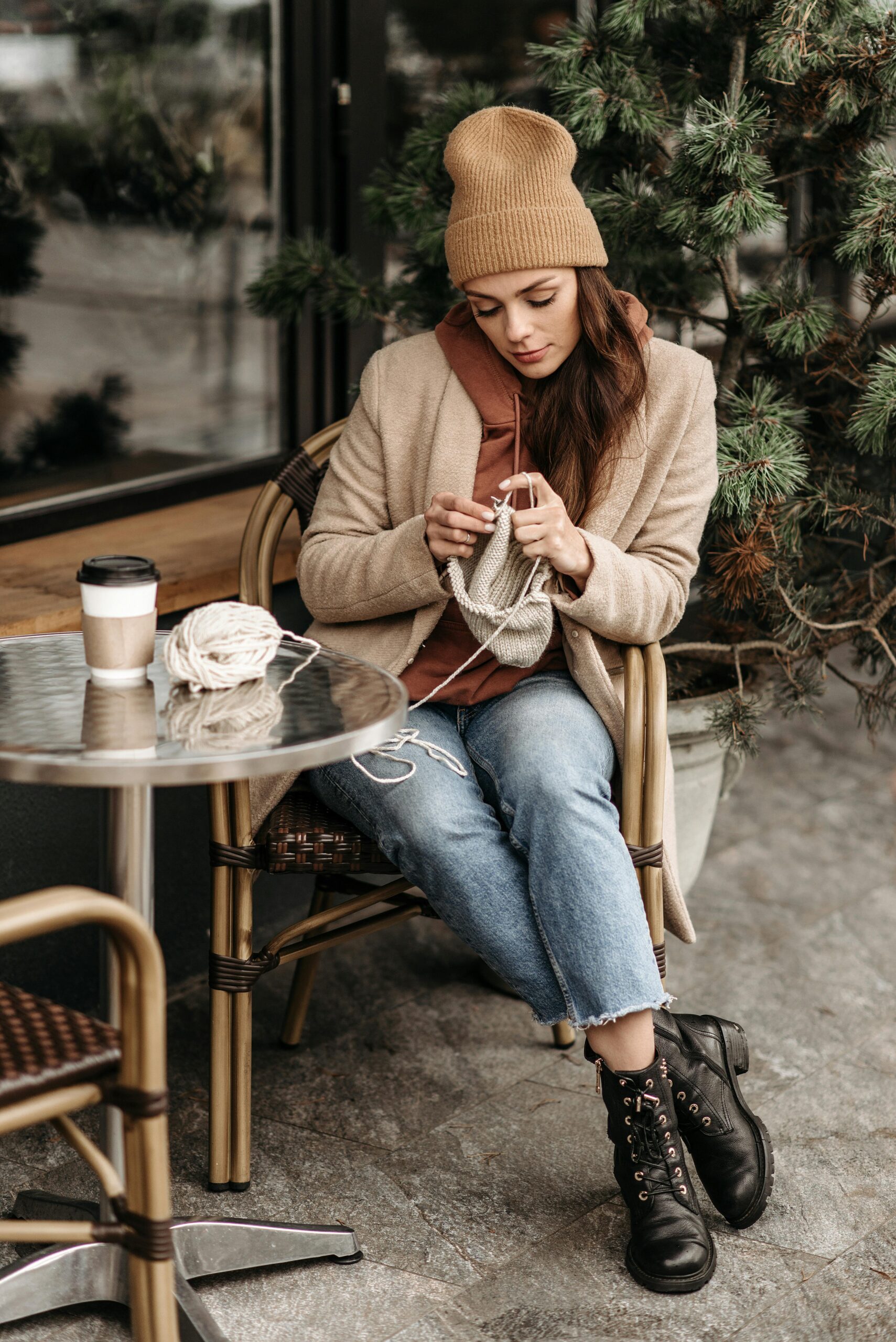 Woman knitting while seated outside a cafe, enjoying leisure time with a hot drink.